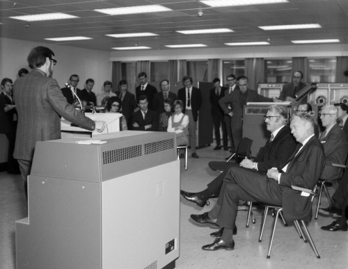 C. H. Devonald fra International Computers Ltd (ICL) står på talerstolen ved overleveringen av Jernverkets nye dataanlegg i 1972. I bakgrunn står de ansatte ved EDB-avdelingen, mens adm. Dir. Per Blidensol sitter i forkant, sammen med blant annet salgsdirektør Å. Laukholm fra Norsk Data og driftsdirektør Sven Edin.